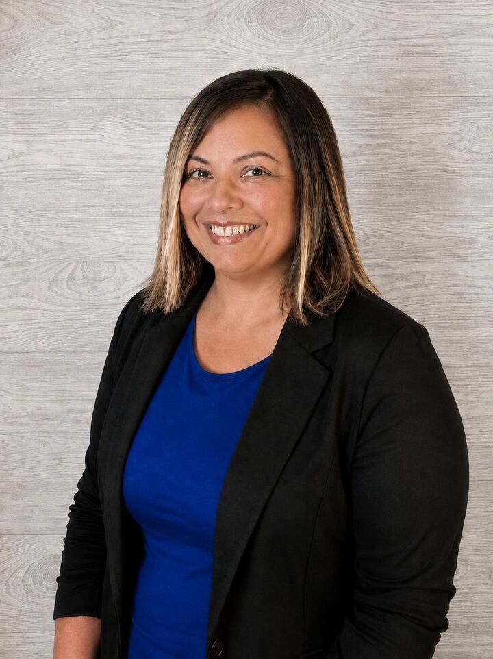 Smiling person in a black blazer and blue top against a light gray textured background