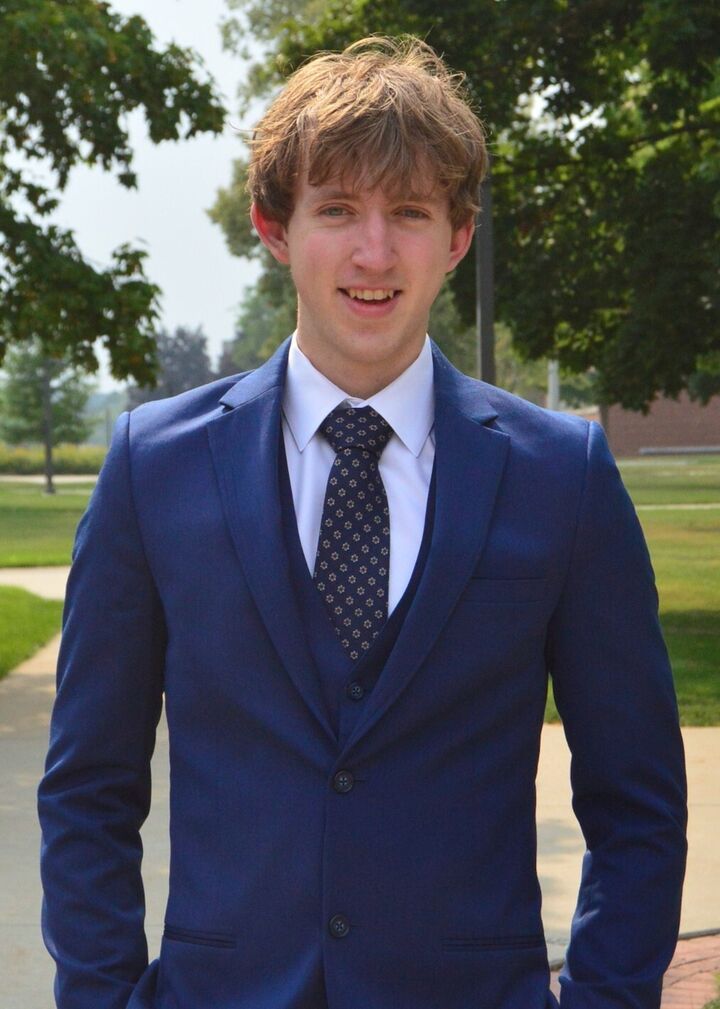 Man in a blue suit and tie standing outdoors on a sunny lawn with trees in the background