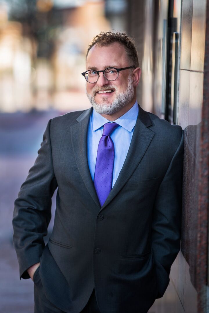Smiling man in a dark suit with blue shirt and purple tie, standing outdoors by a wall.