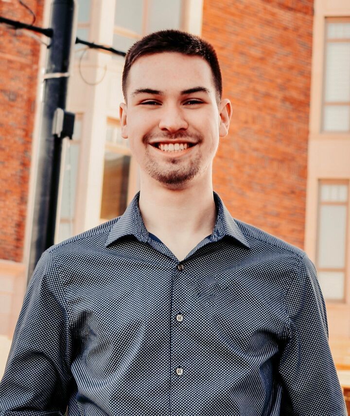 Smiling man in a dark patterned shirt standing outdoors in front of a brick wall
