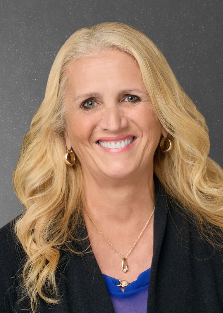 Smiling blonde woman in a black blazer and blue top against a gray background