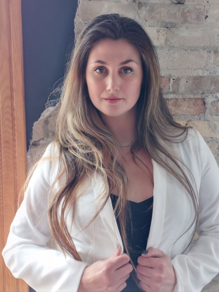 Woman in a white blazer standing against a brick wall, holding her lapels with a neutral expression.