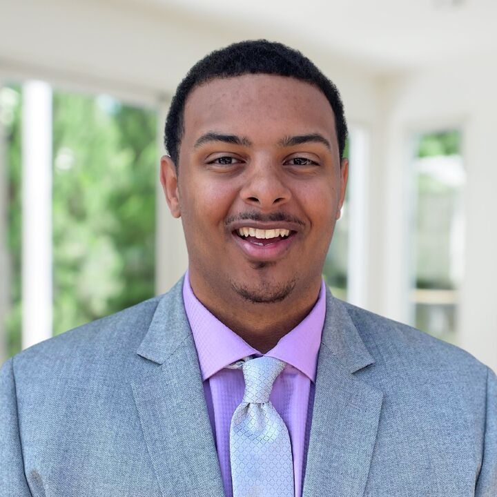 Portrait of a smiling person in a light gray suit and lavender shirt outdoors