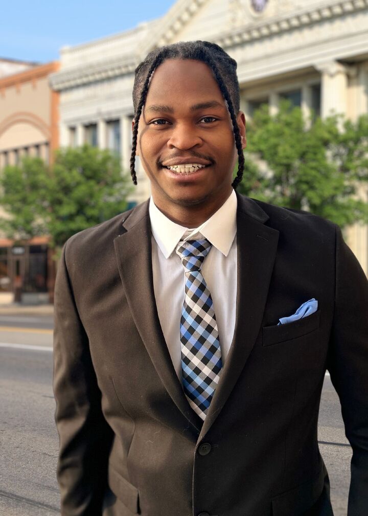 Smiling man in a dark suit and plaid tie standing outdoors in front of a building