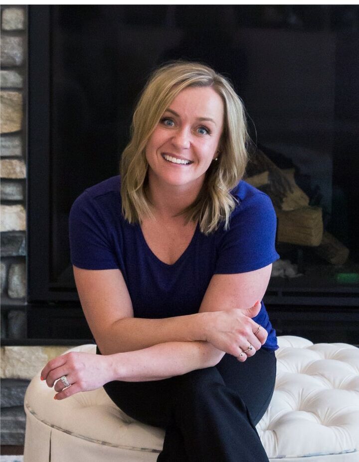 Smiling woman in a navy shirt sitting on a white tufted ottoman in front of a dark fireplace.