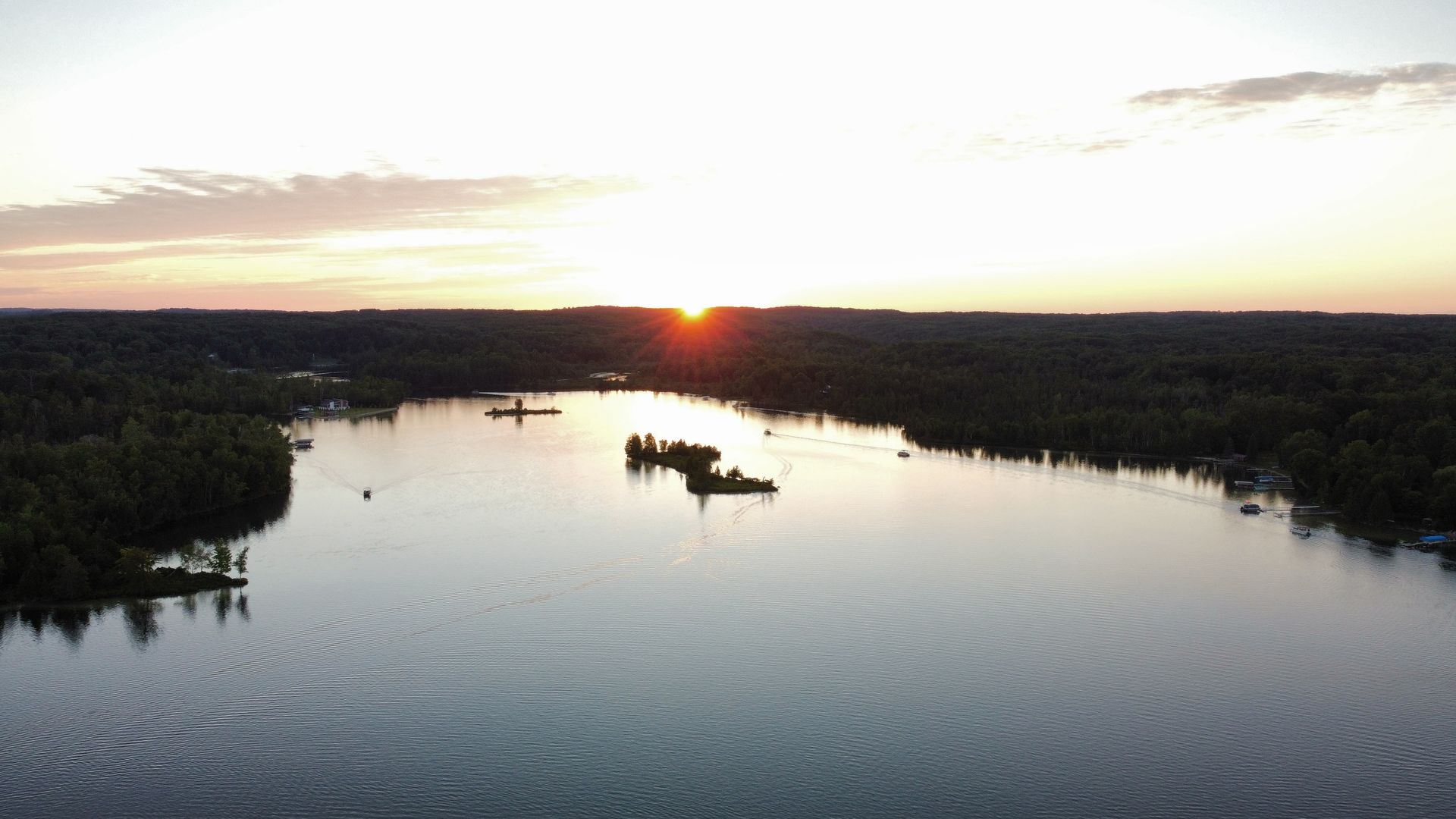 Sunset over a calm river bordered by dark forest, with small boats and a glowing horizon
