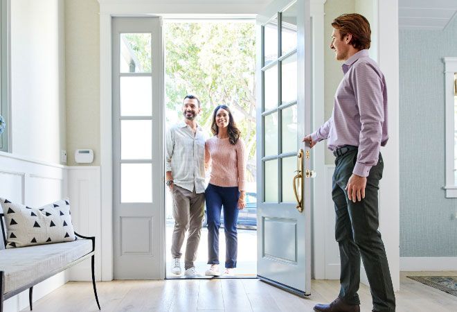 Two people entering a bright home while a man greets them at the open front door.