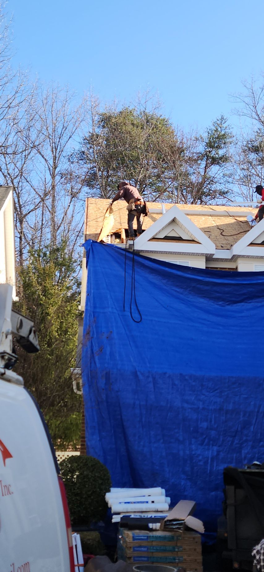 A man is working on the roof of a house behind a blue tarp.