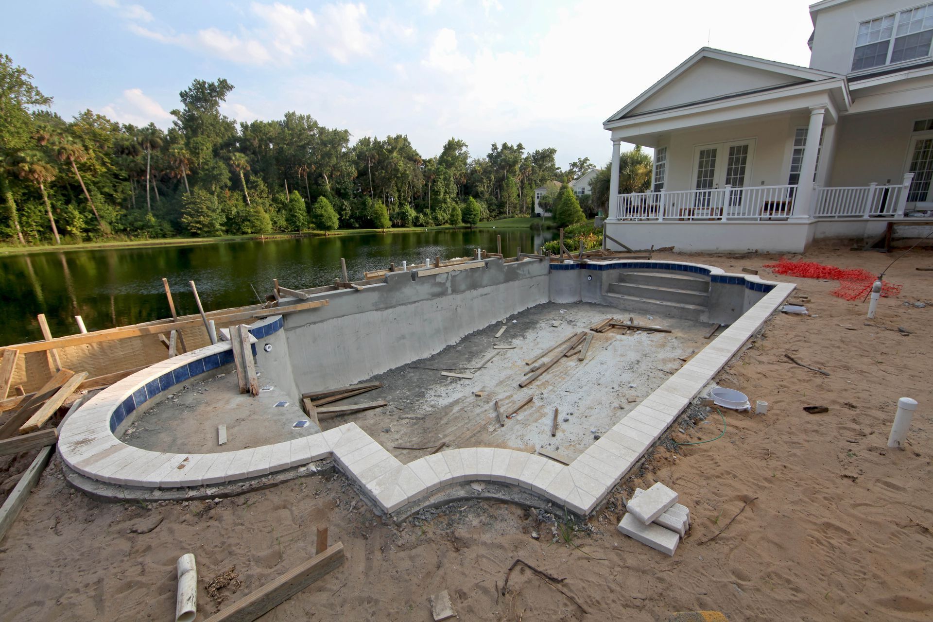 Partially built in-ground pool beside a house, with sandy yard and trees by the water