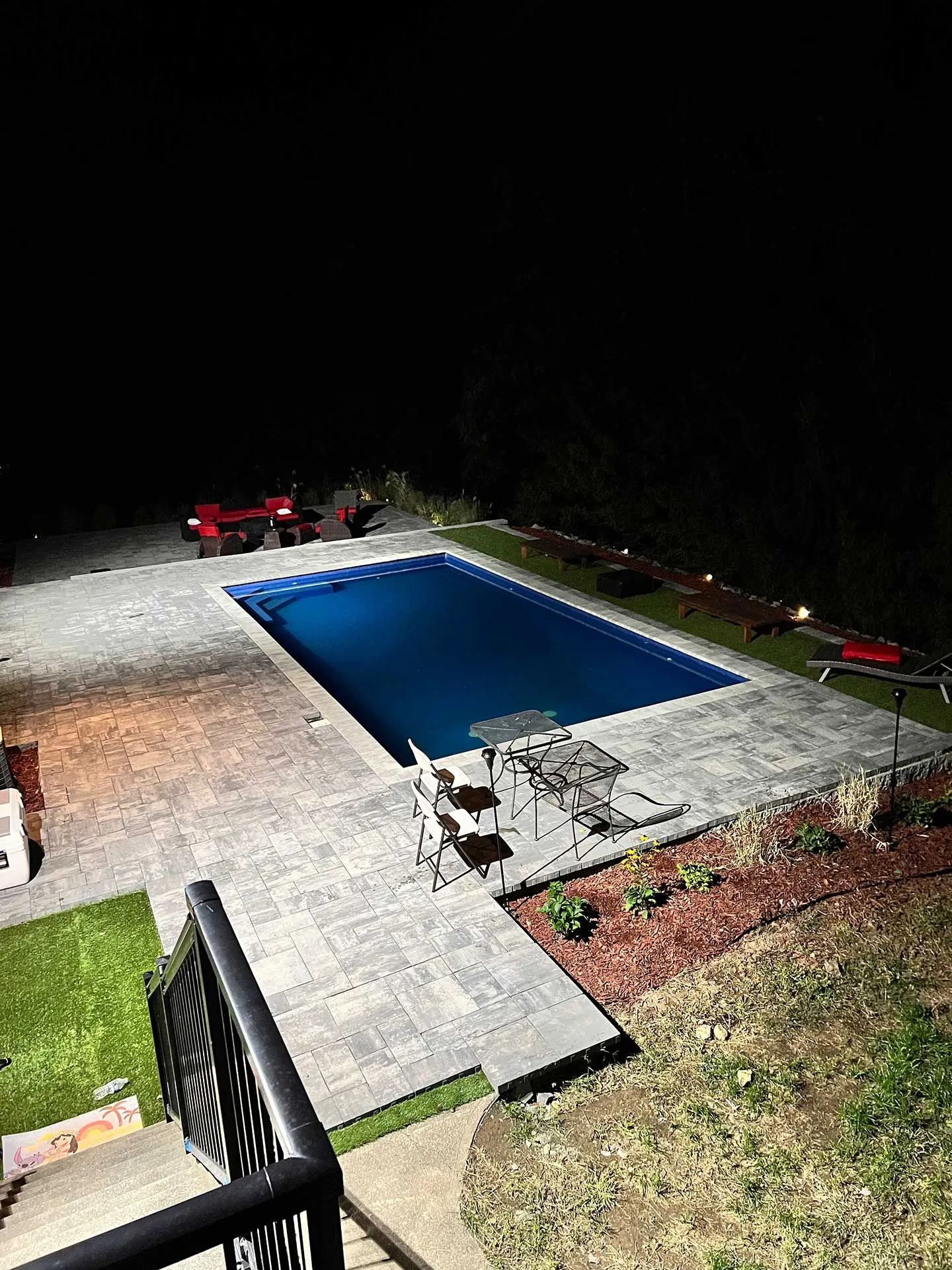 Backyard pool lit at night, surrounded by stone patio and lawn