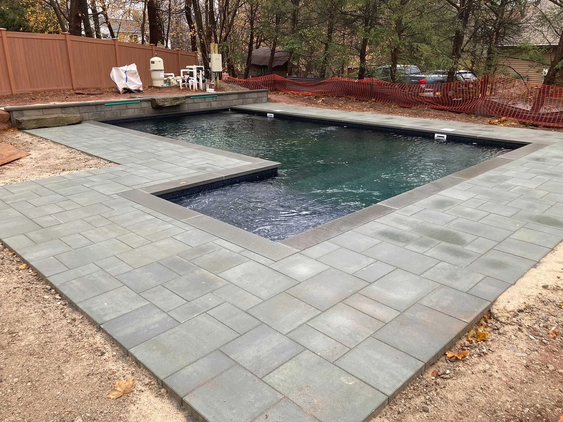 Backyard swimming pool with gray paver patio and a raised spa, surrounded by fencing and trees