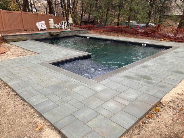 Backyard swimming pool with gray stone deck and red safety fence, surrounded by trees