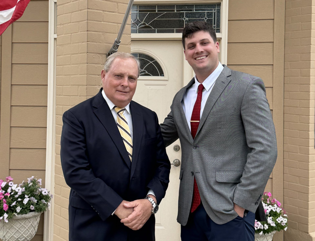 Two men in suits standing by a house entrance with an American flag and flower planters.