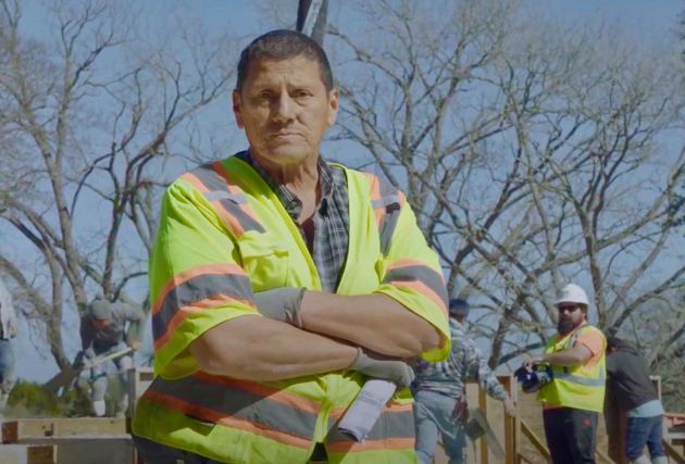 A foreman in a yellow safety vest stands with arms crossed at a construction site while workers build in the background.