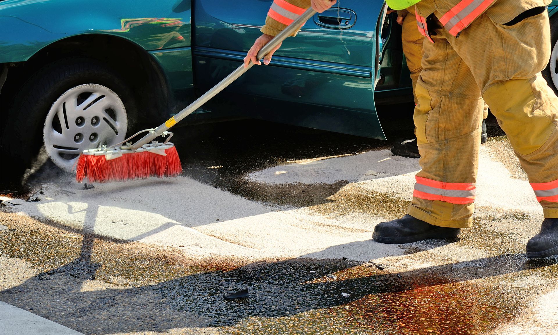 A firefighter in protective gear uses a broom to scrub a spill on the asphalt near a car wheel.