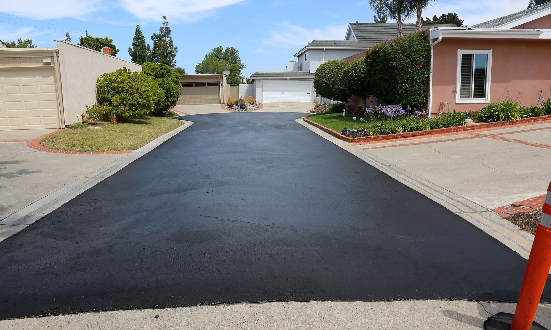 A freshly paved, dark asphalt driveway between two suburban houses with lawns and a traffic cone on the right.