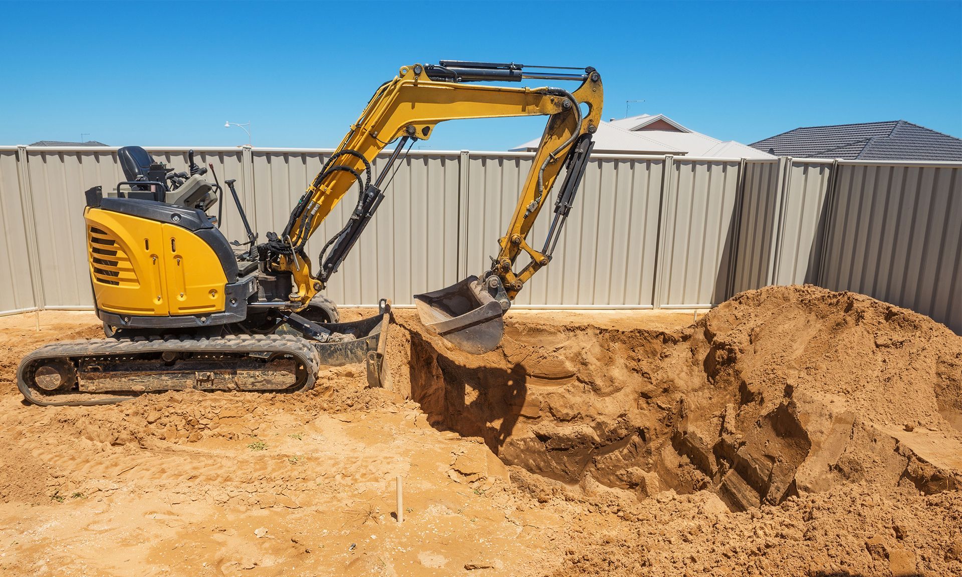 A yellow compact excavator digs a hole in the sandy ground of a residential backyard with a fence in the background.