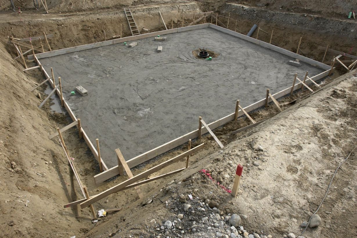 A rectangular concrete foundation poured within a wooden formwork inside an excavated dirt construction site.