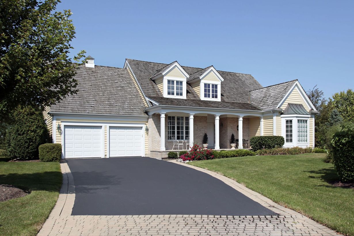 A cream-colored suburban house with a two-car garage, a covered front porch, and a paved driveway.