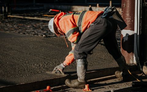 A construction worker in a high-visibility orange jacket and white hard hat uses a hand tool to smooth wet concrete.