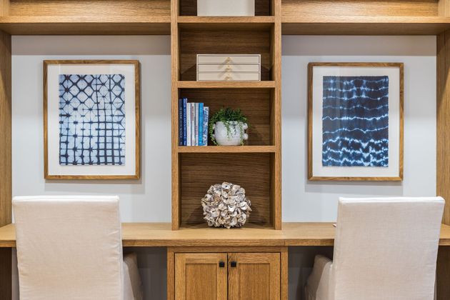 A home office desk with two white chairs, wooden shelving, and two blue patterned framed prints on a white wall.