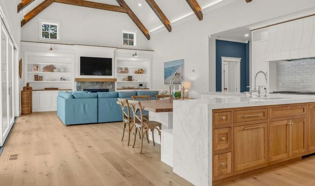 A bright open-concept living area with wood beams, a light blue sofa, a marble kitchen island, and natural wood cabinetry.