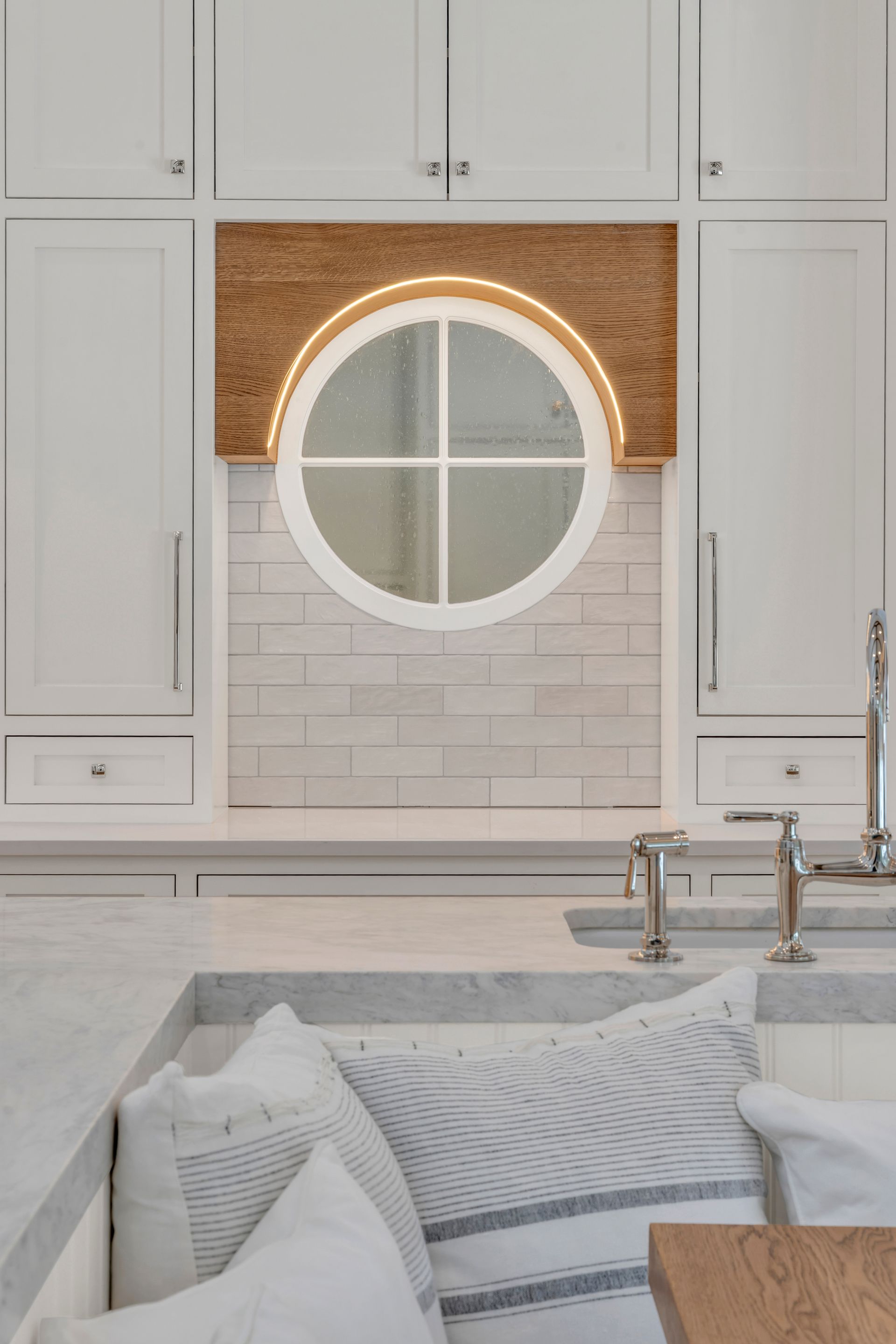 A circular window framed by wood in a white kitchen above a counter with gray stone and patterned throw pillows.