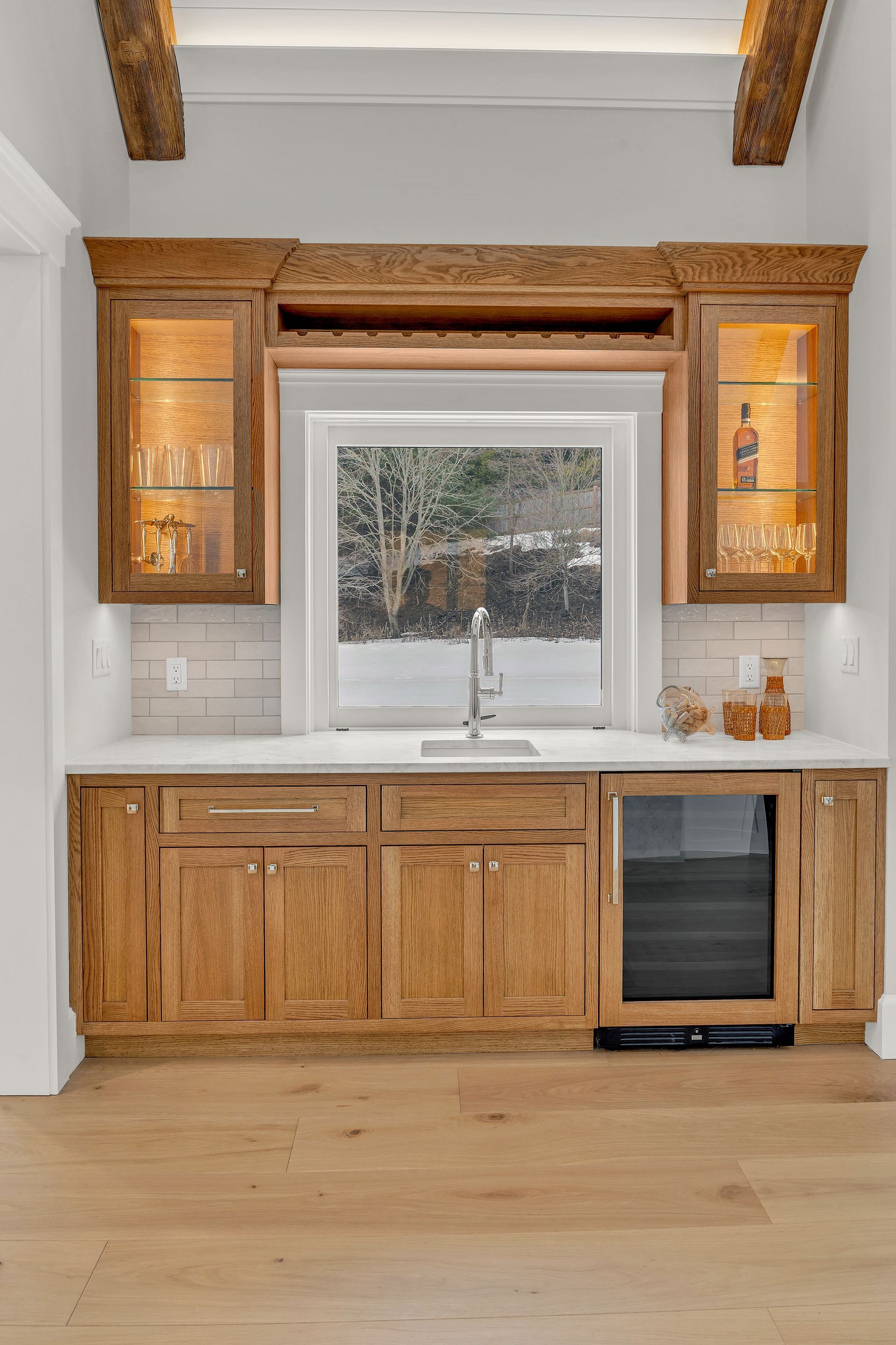 A wet bar with rustic wood cabinets, a sink, glass-front upper cabinets, and a wine fridge against a white wall.