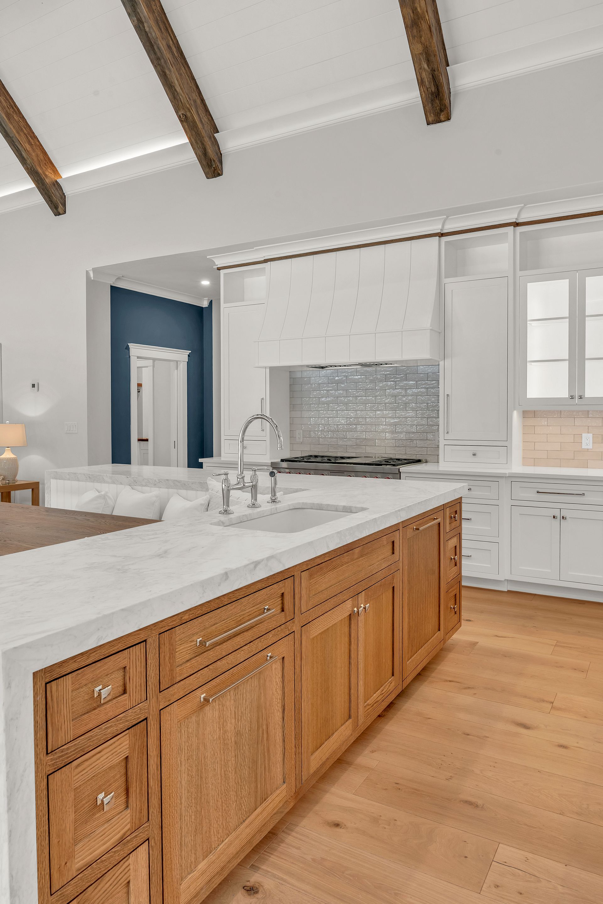 A bright kitchen featuring a large wooden island with a white countertop and white cabinets, under exposed ceiling beams.