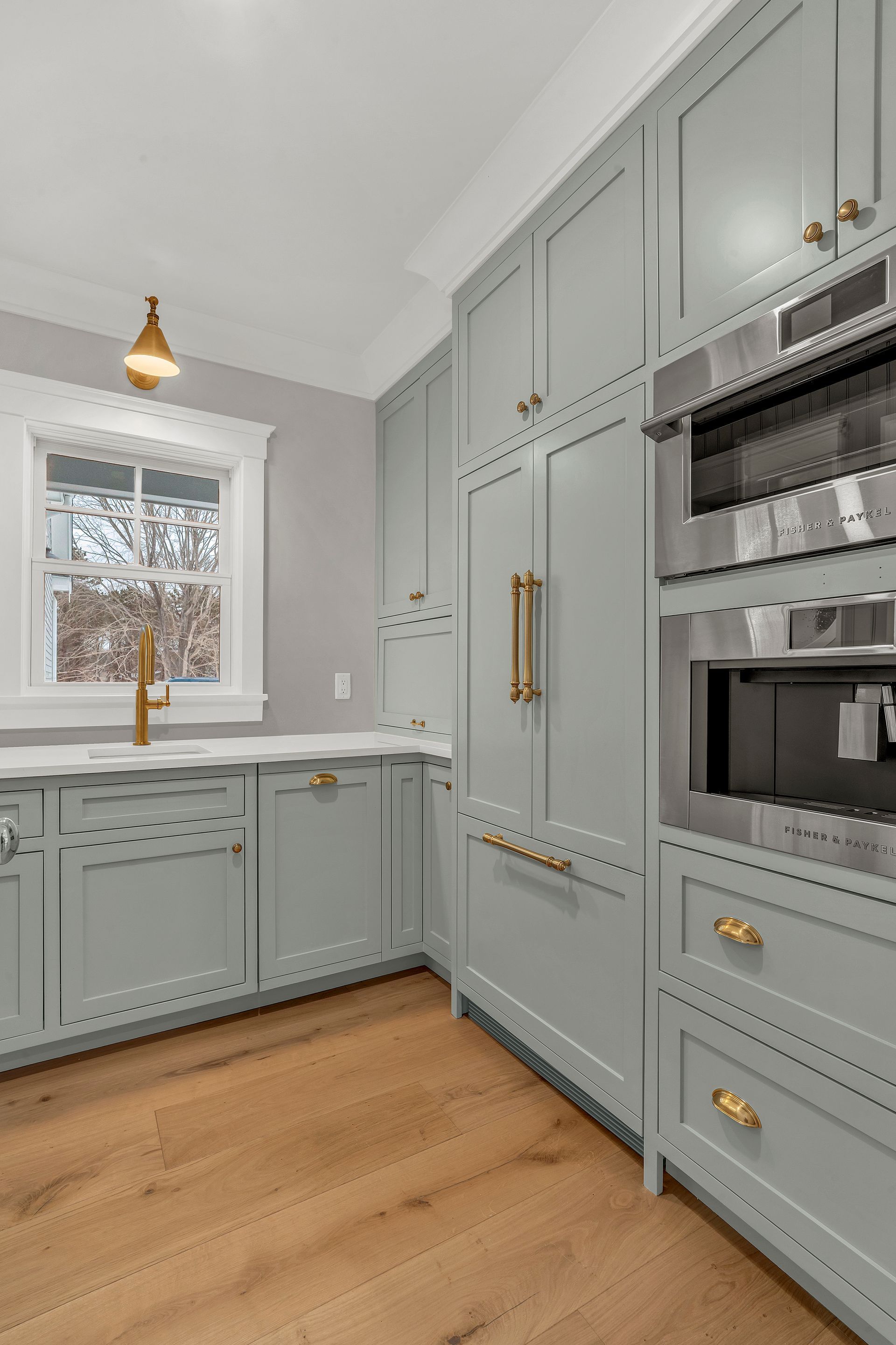 A kitchen with sage green cabinets, light wood floors, a window with a brass faucet, and built-in stainless appliances.