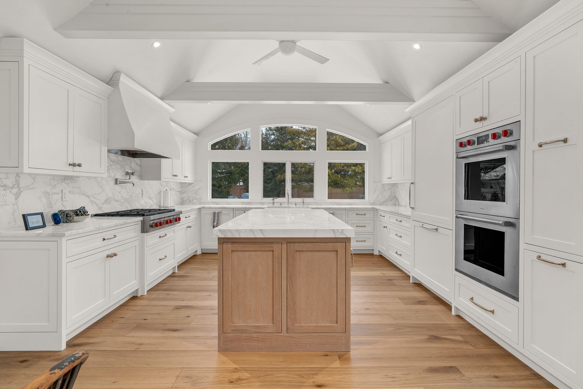A symmetrical white kitchen with wooden floors, a large central island, and double ovens under a vaulted ceiling.