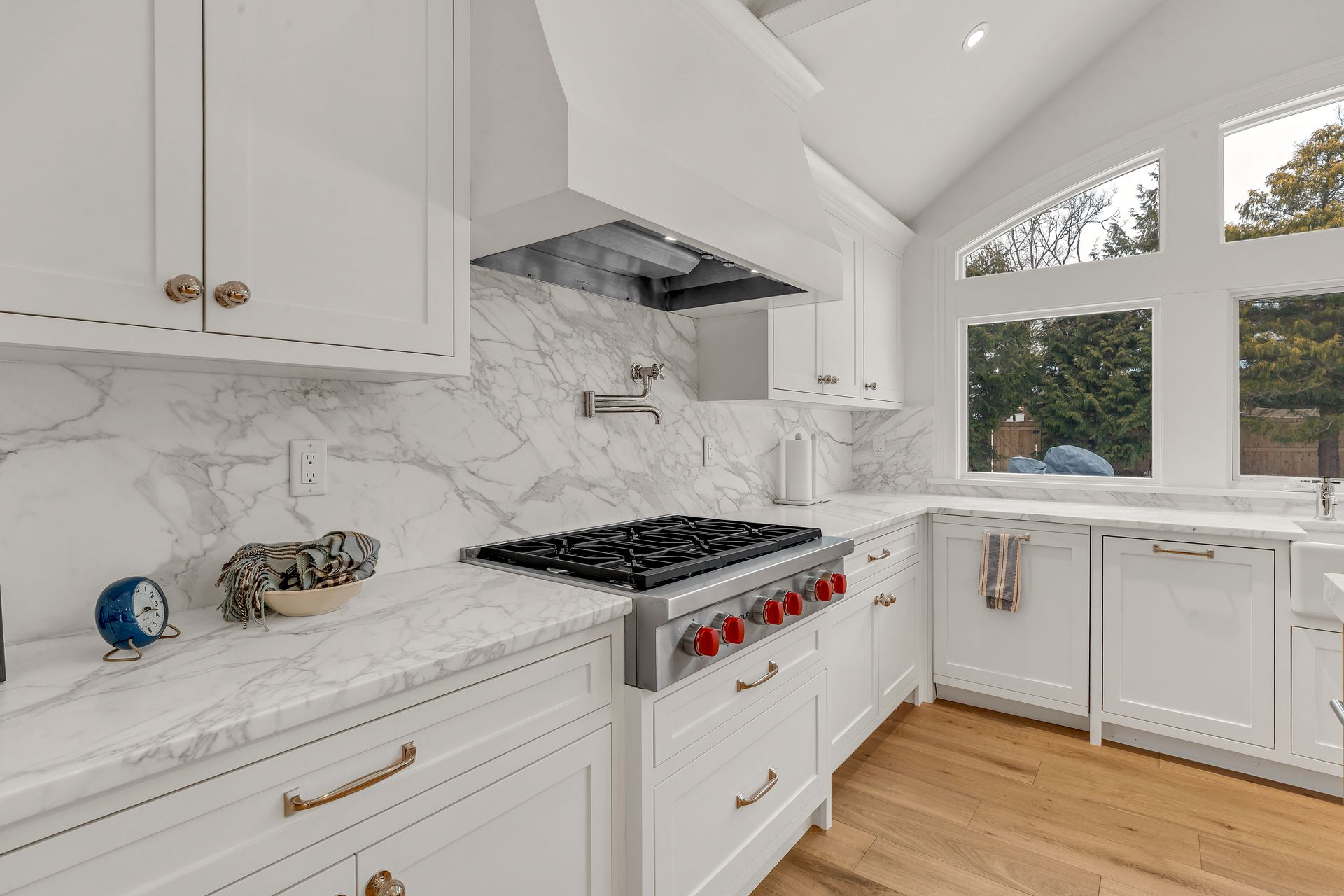 A bright, modern kitchen featuring white cabinets, marble countertops and backsplash, a gas range, and wood flooring.