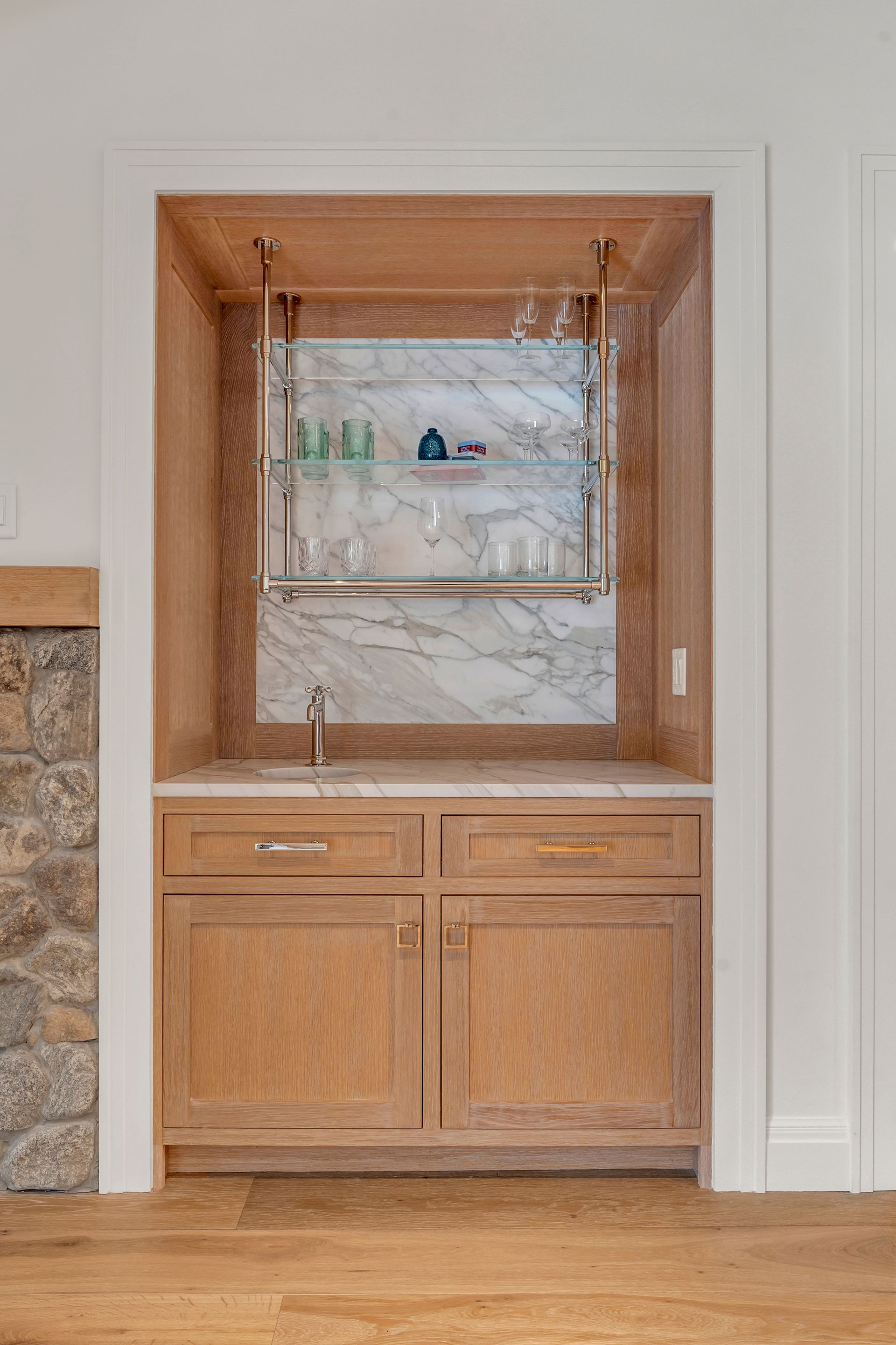 A built-in wet bar with light wood cabinetry, a white marble countertop and backsplash, and a suspended glass shelf.