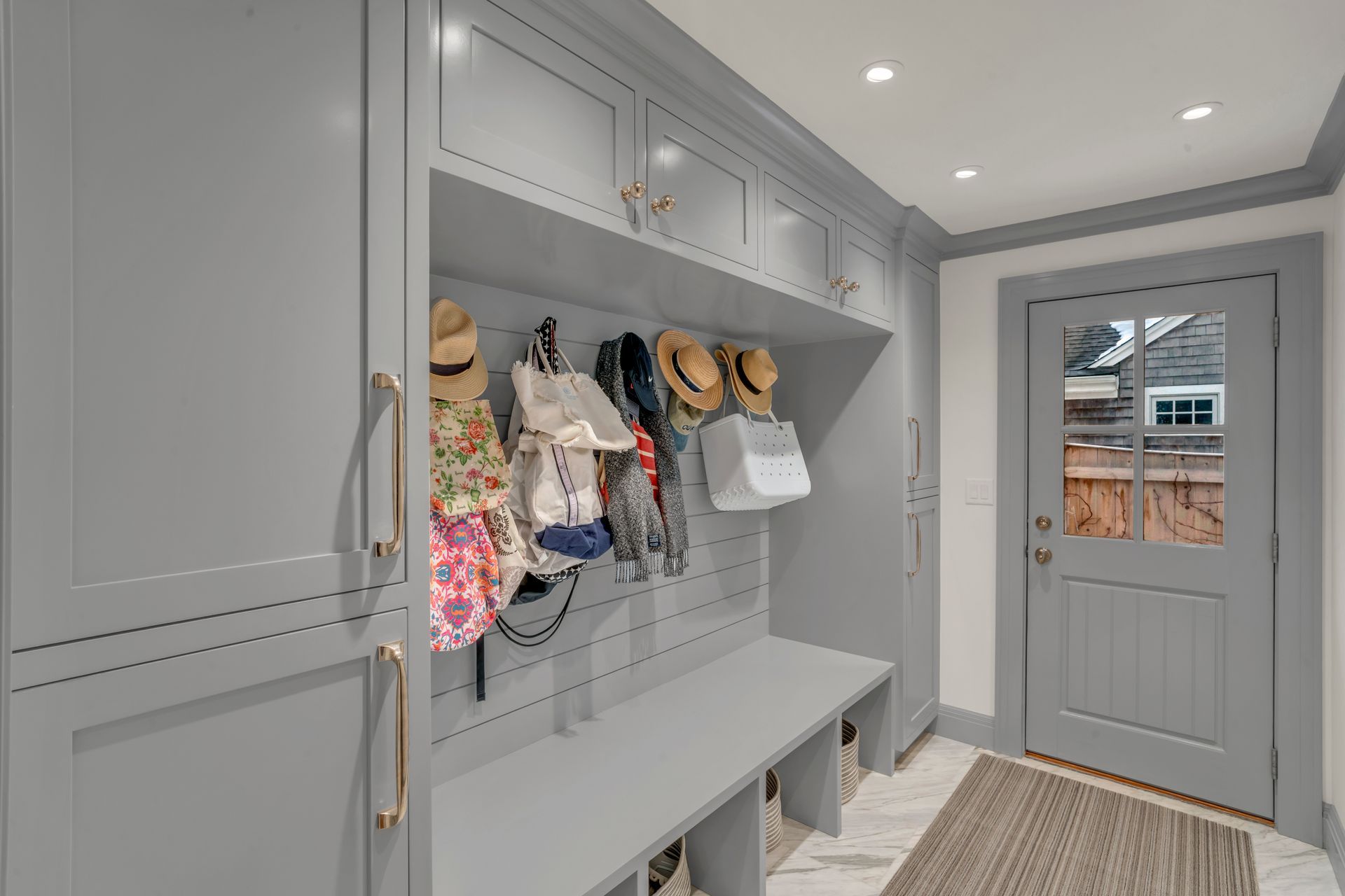 A grey mudroom entry with built-in cabinetry, a bench, coat hooks with bags and hats, and a glass-paneled door.