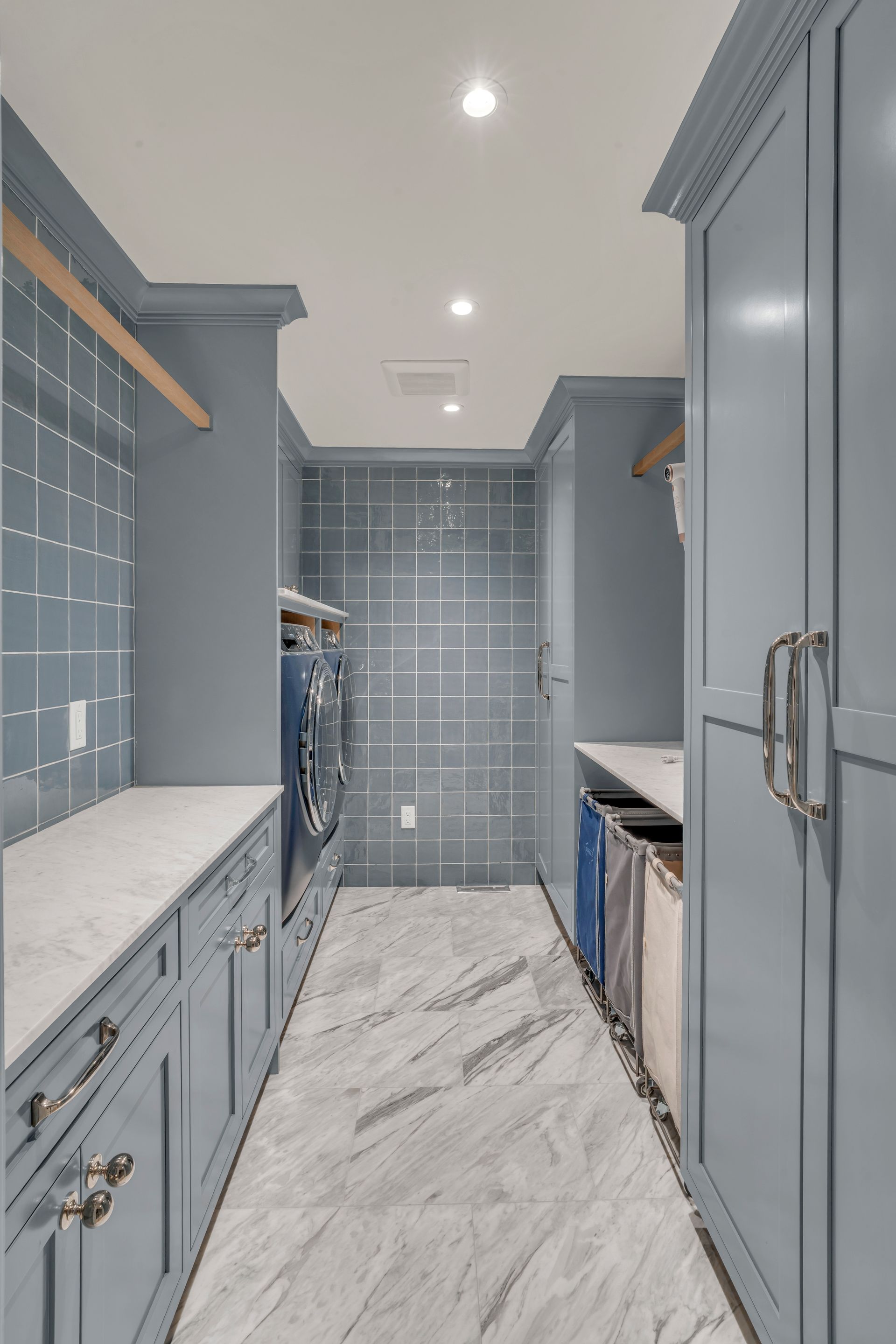 A modern laundry room with blue cabinets, marble countertops, tiled walls, and white marble-patterned flooring.