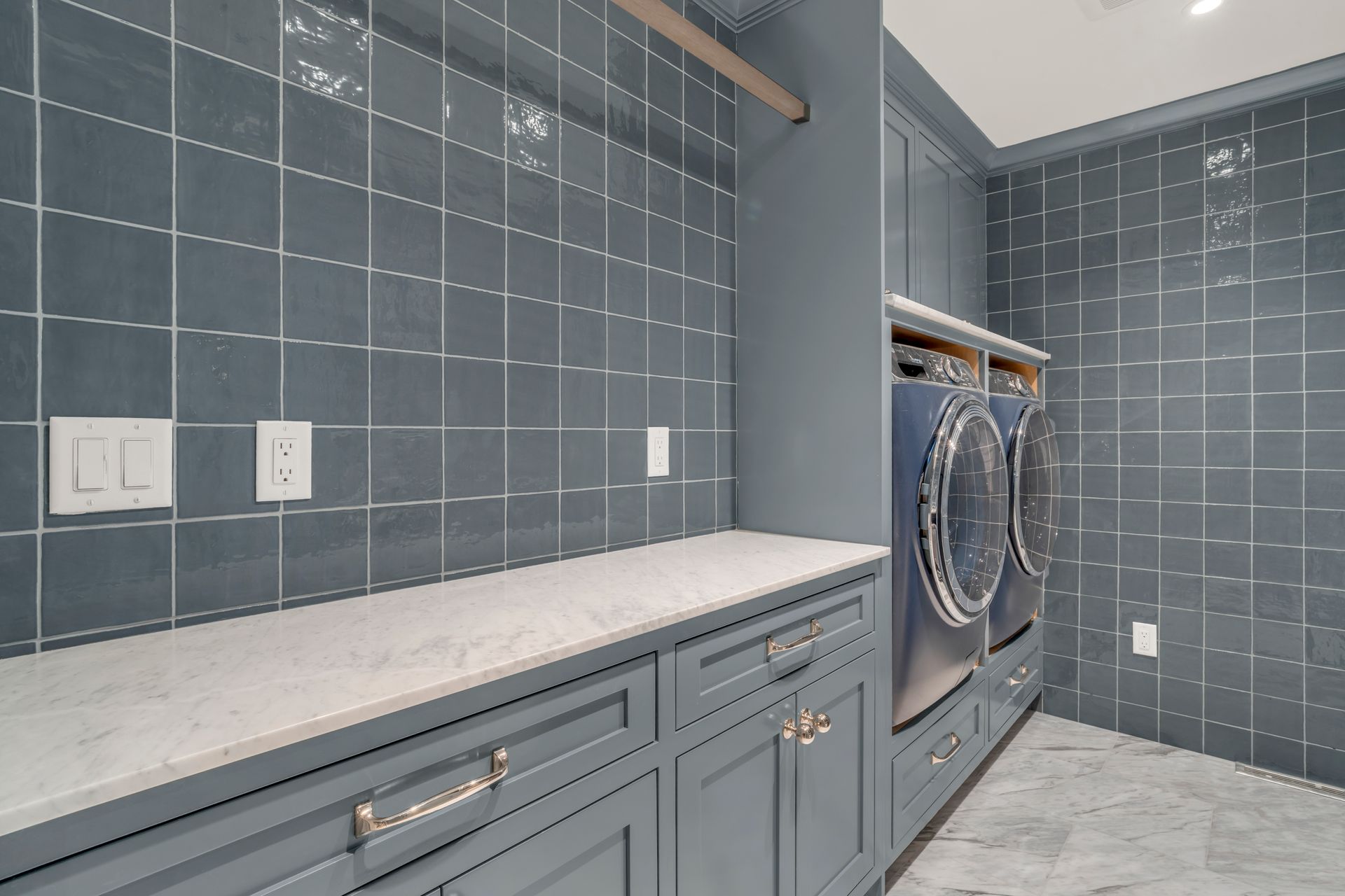 Blue-gray laundry room with tiled walls, marble countertops, dark cabinetry, and a stacked washer and dryer unit.
