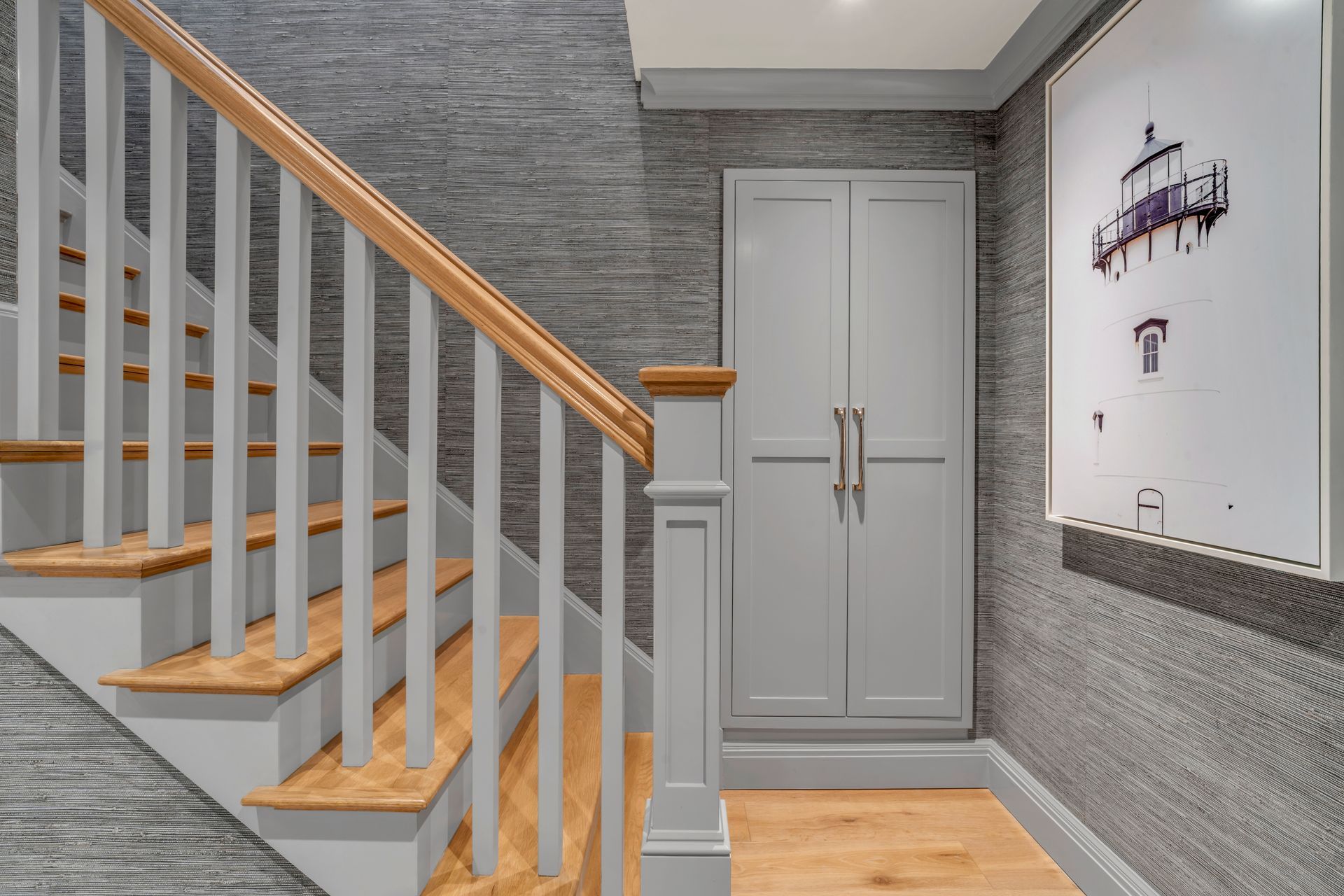 Staircase with a wooden handrail and light-colored spindles next to a tall, grey cabinet against a textured grey wall.