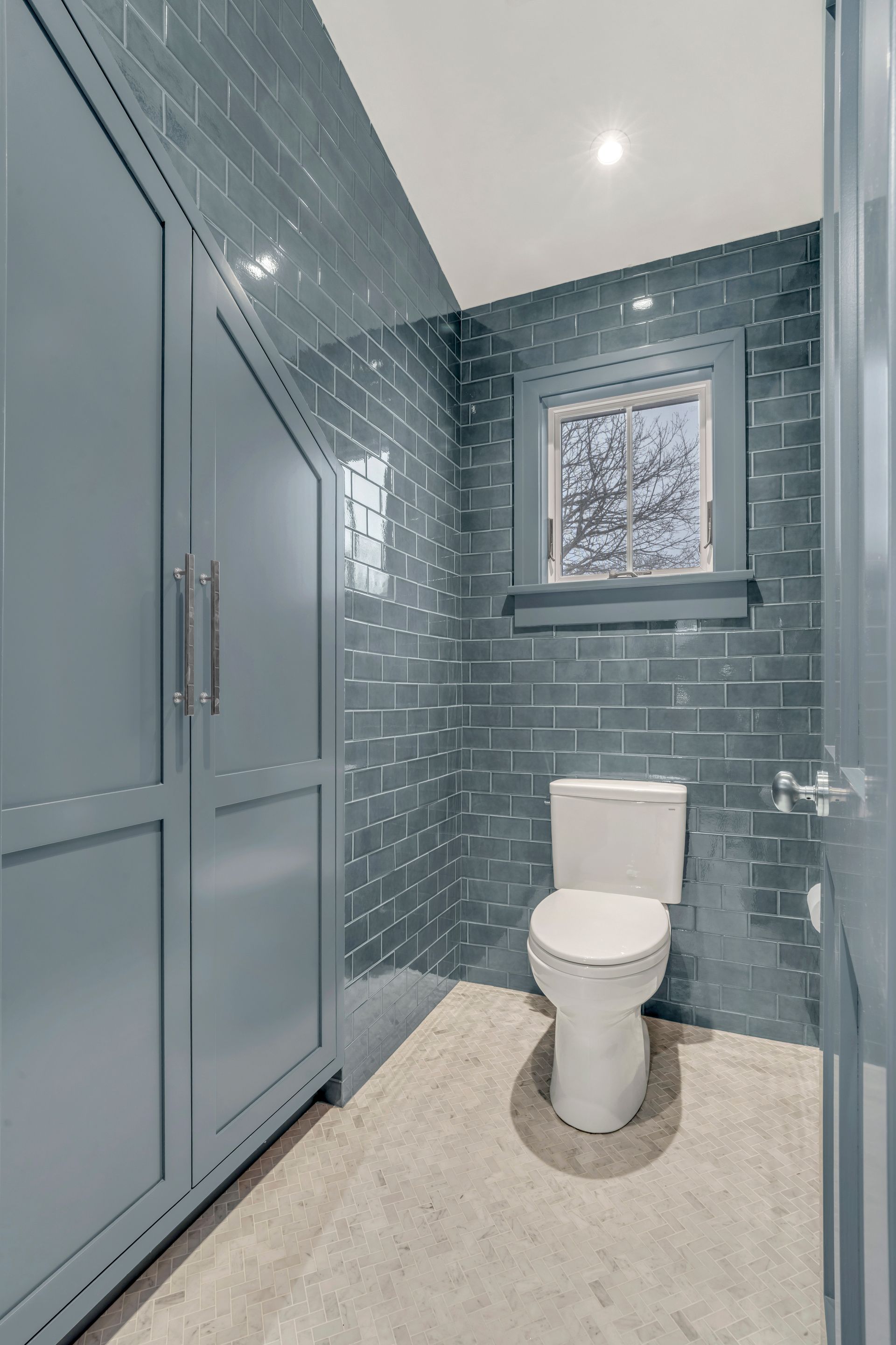 A bathroom with dark blue, glossy subway tiles, a white toilet, and a custom blue storage cabinet with slanted top doors.