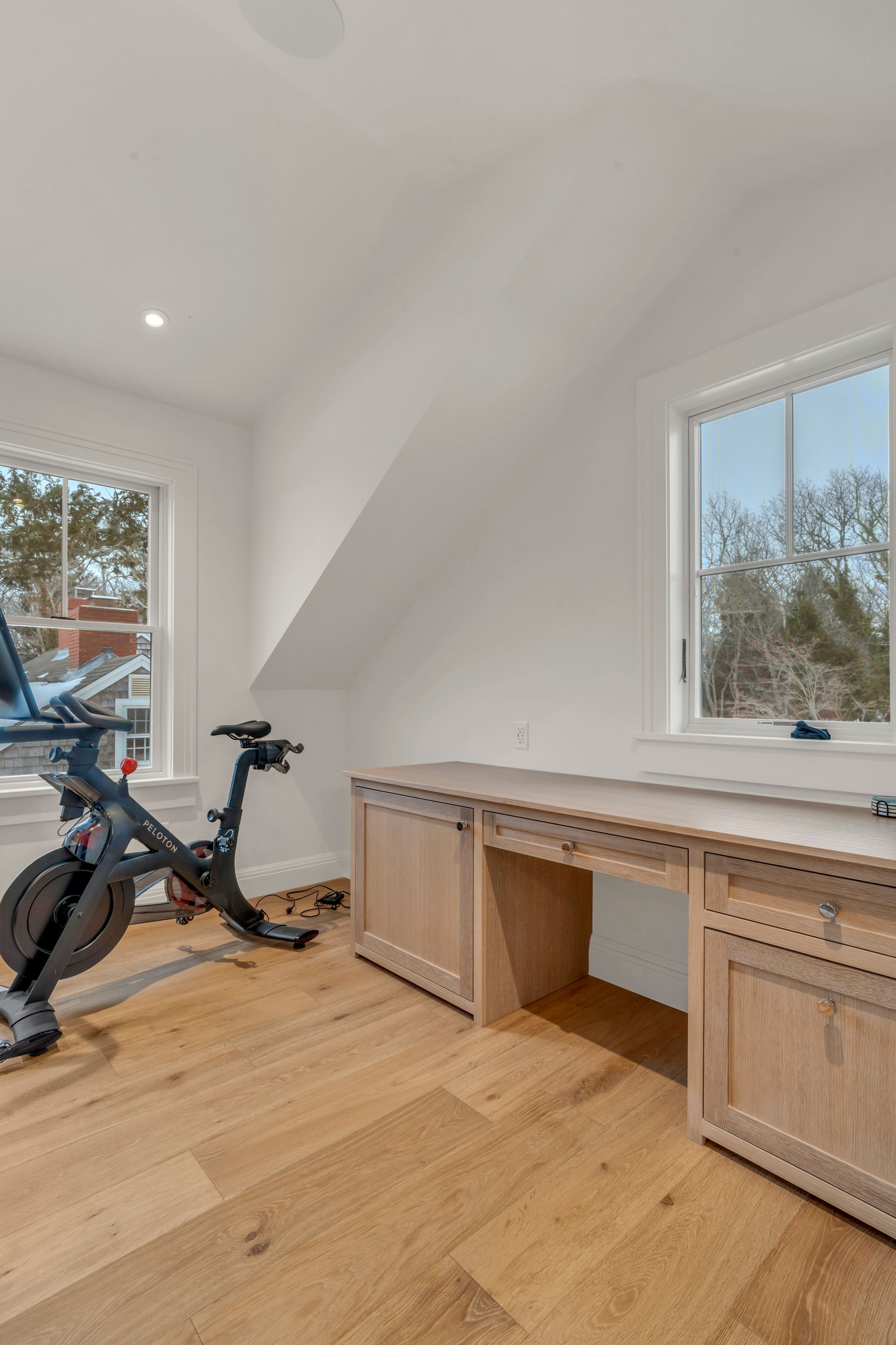 A room with a stationary bike next to a light wood desk featuring drawers and cabinets beneath a window.