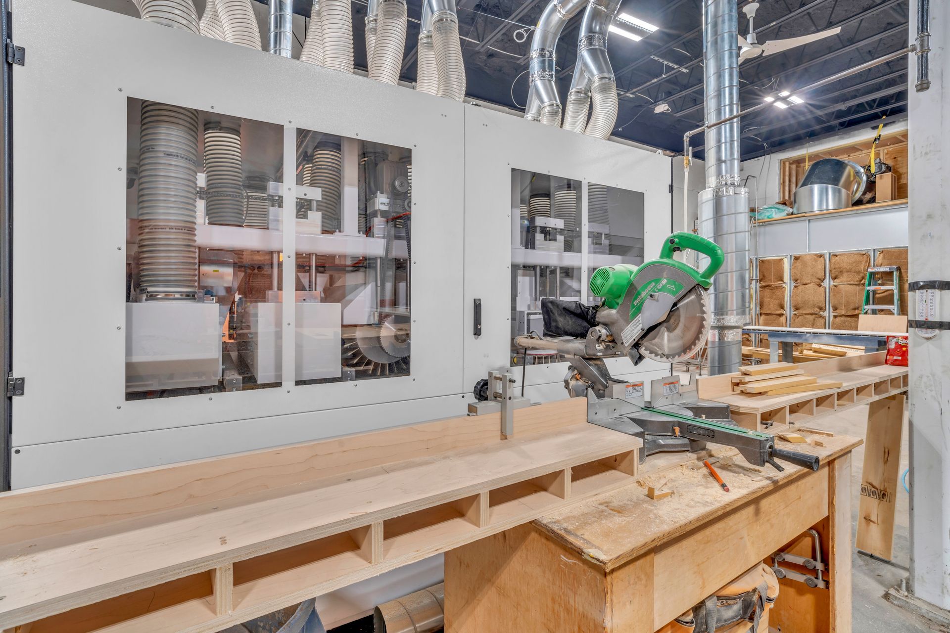 A green miter saw sits on a wooden workbench in a workshop with industrial ventilation pipes and wooden structures.