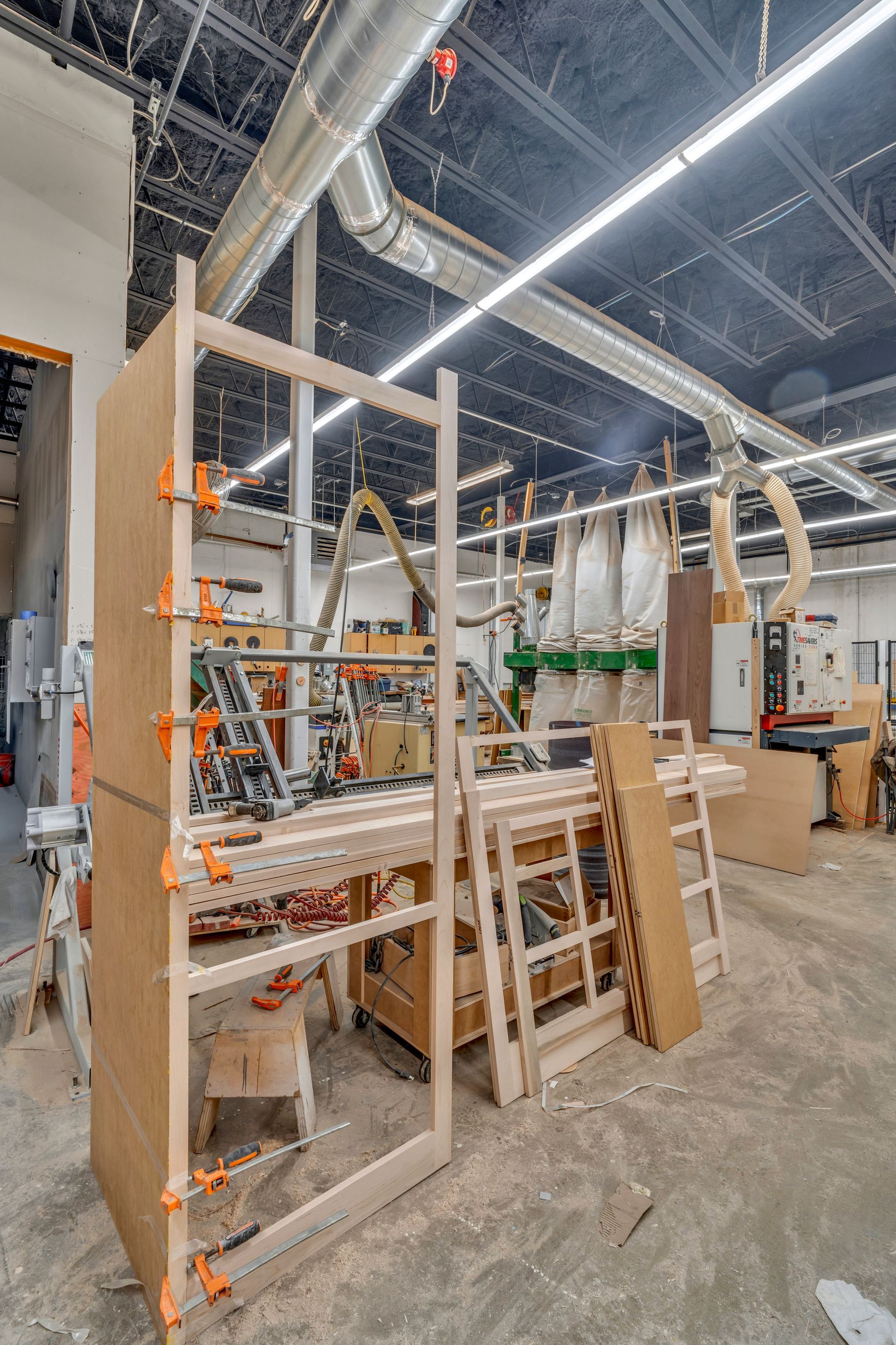 Woodworking shop interior featuring a large wooden frame being assembled with clamps and industrial equipment nearby.
