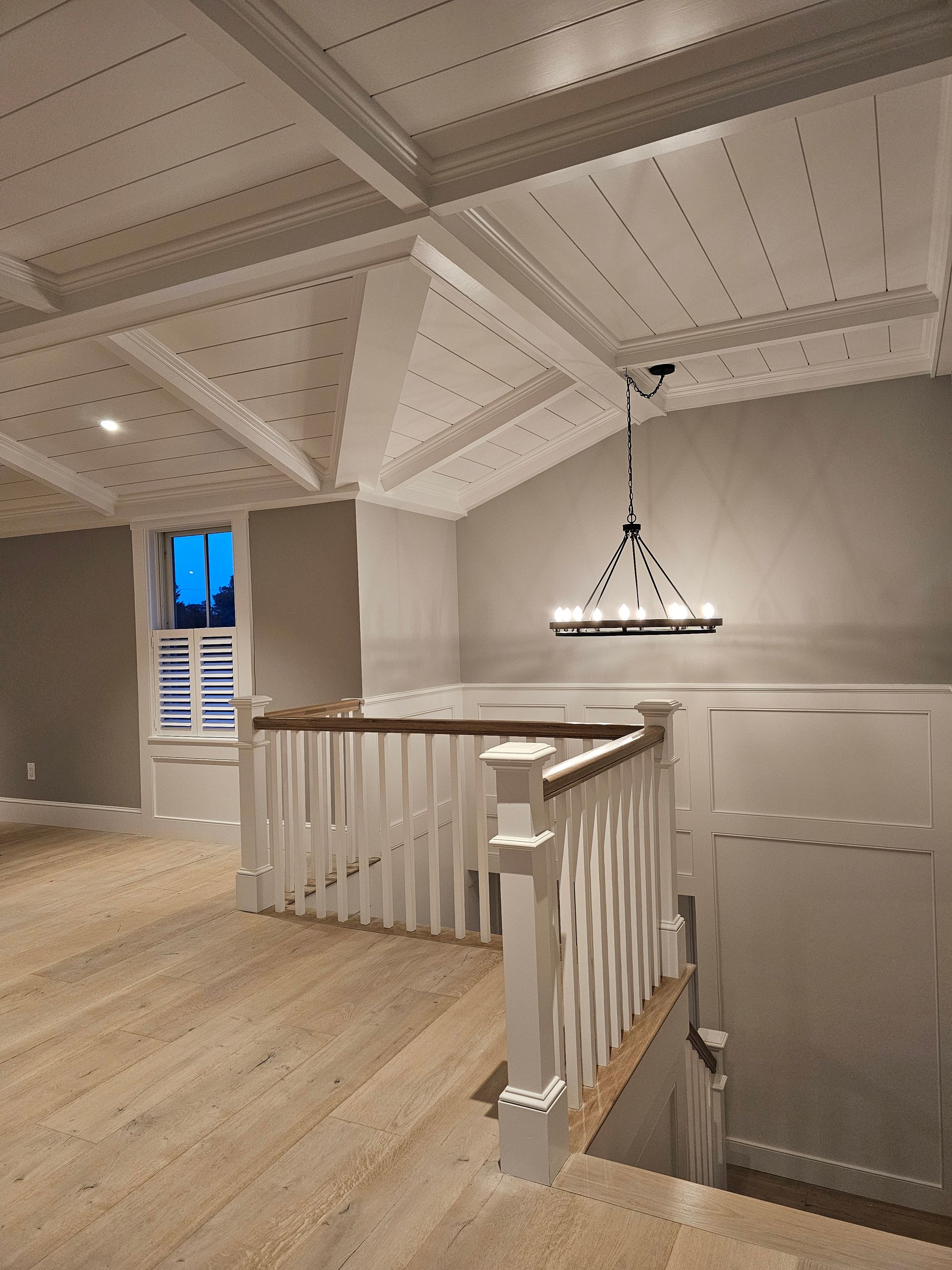 An interior view of a landing area with light wood floors, white paneled ceilings, a wooden staircase, and a chandelier.
