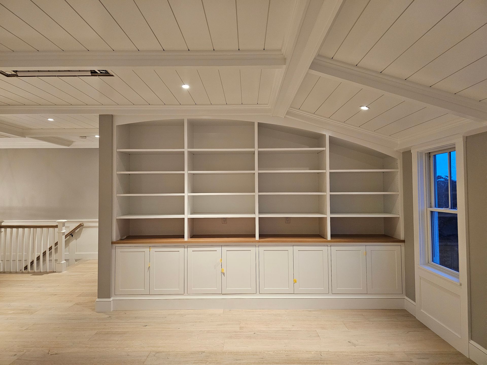 Built-in white bookshelves and cabinets under a slanted, planked ceiling in a room with light wood floors.