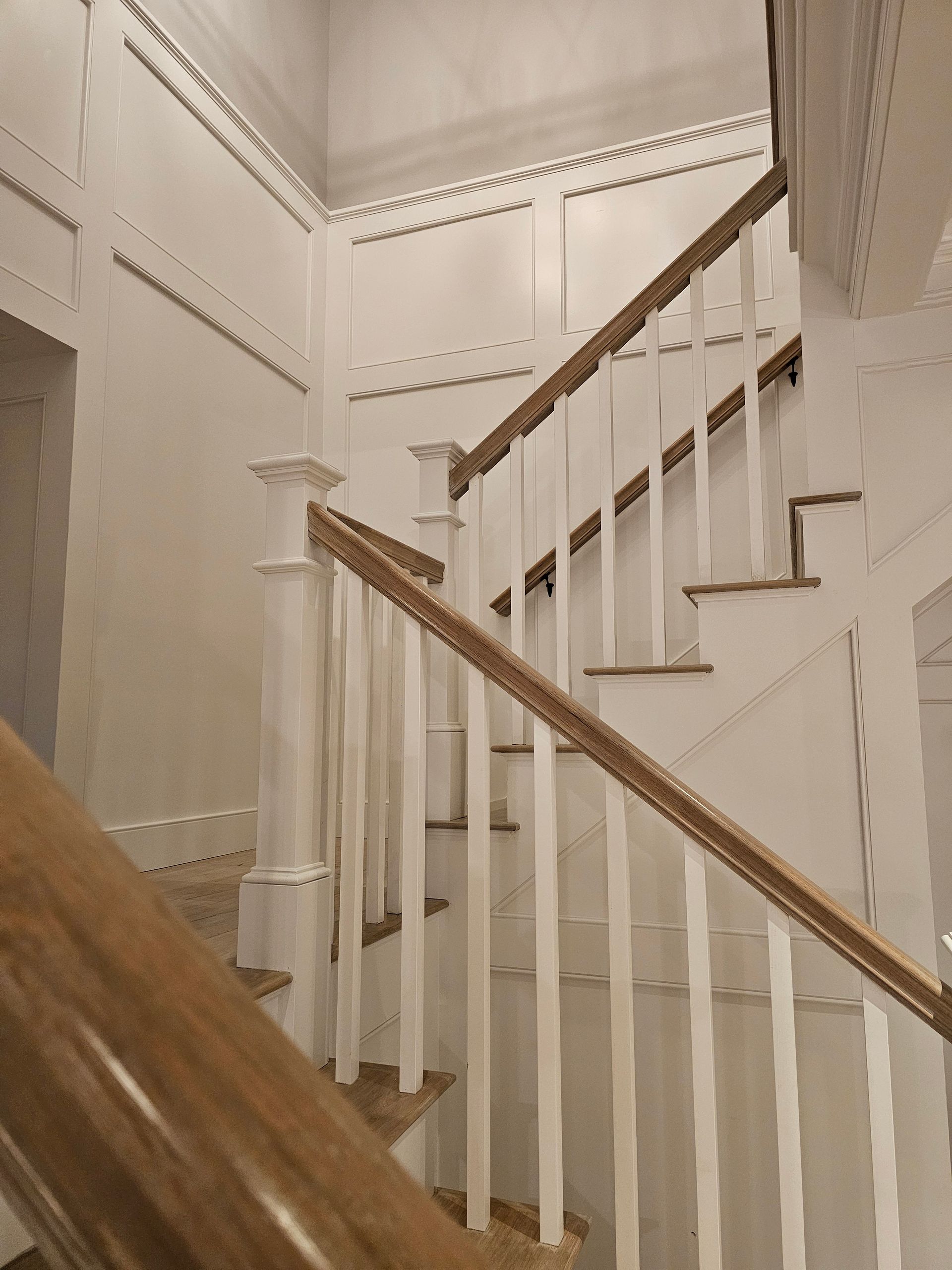 A staircase with white panelled walls, white spindles, and a natural wood handrail, angled upward in a residential interior.