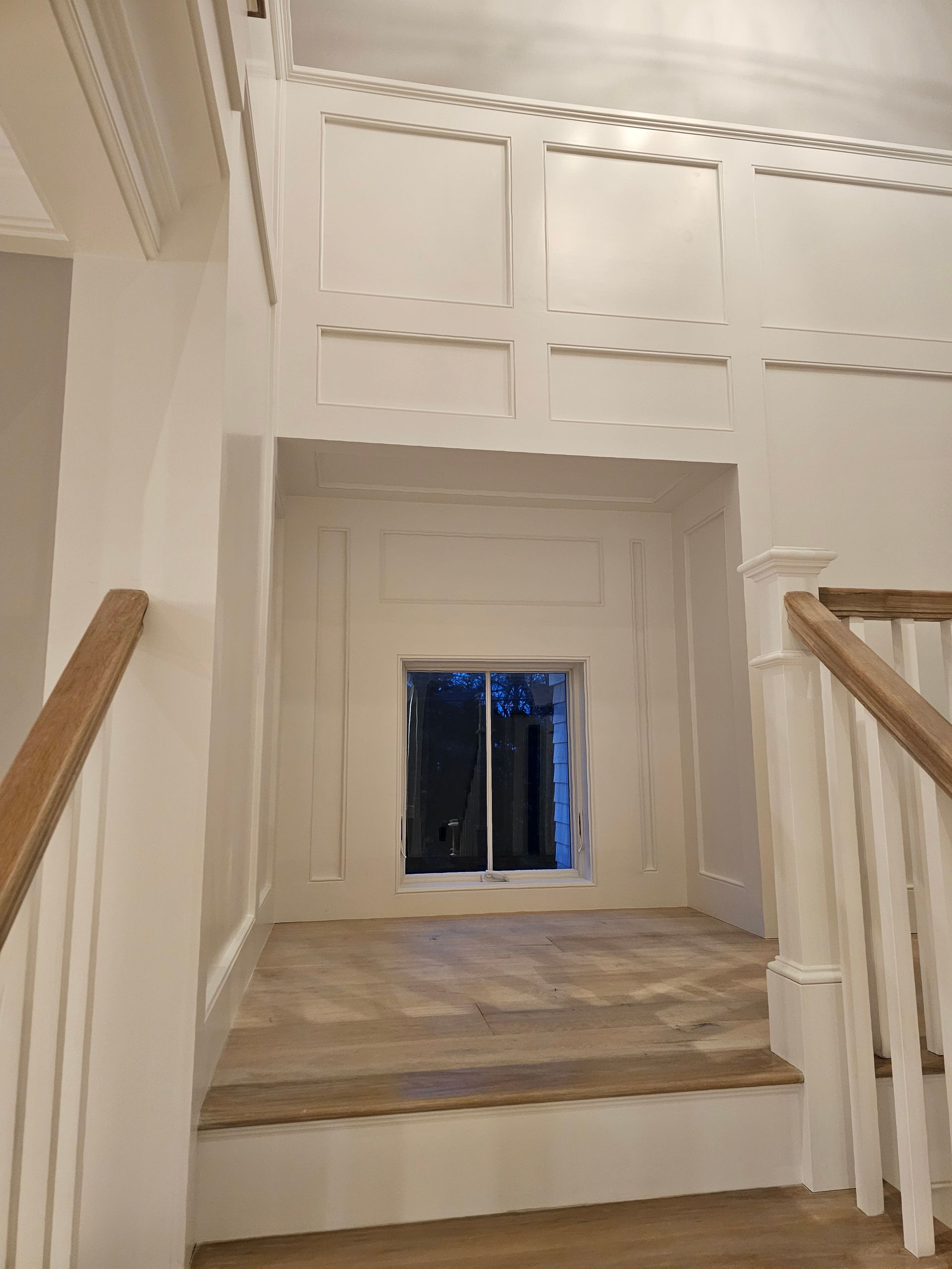 A view looking up a staircase toward a landing with a small window, white paneled walls, and wood flooring.