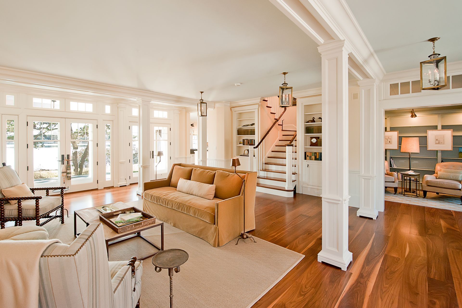 A bright, open-plan living room with beige sofas, wood floors, and a staircase, leading into a study area.
