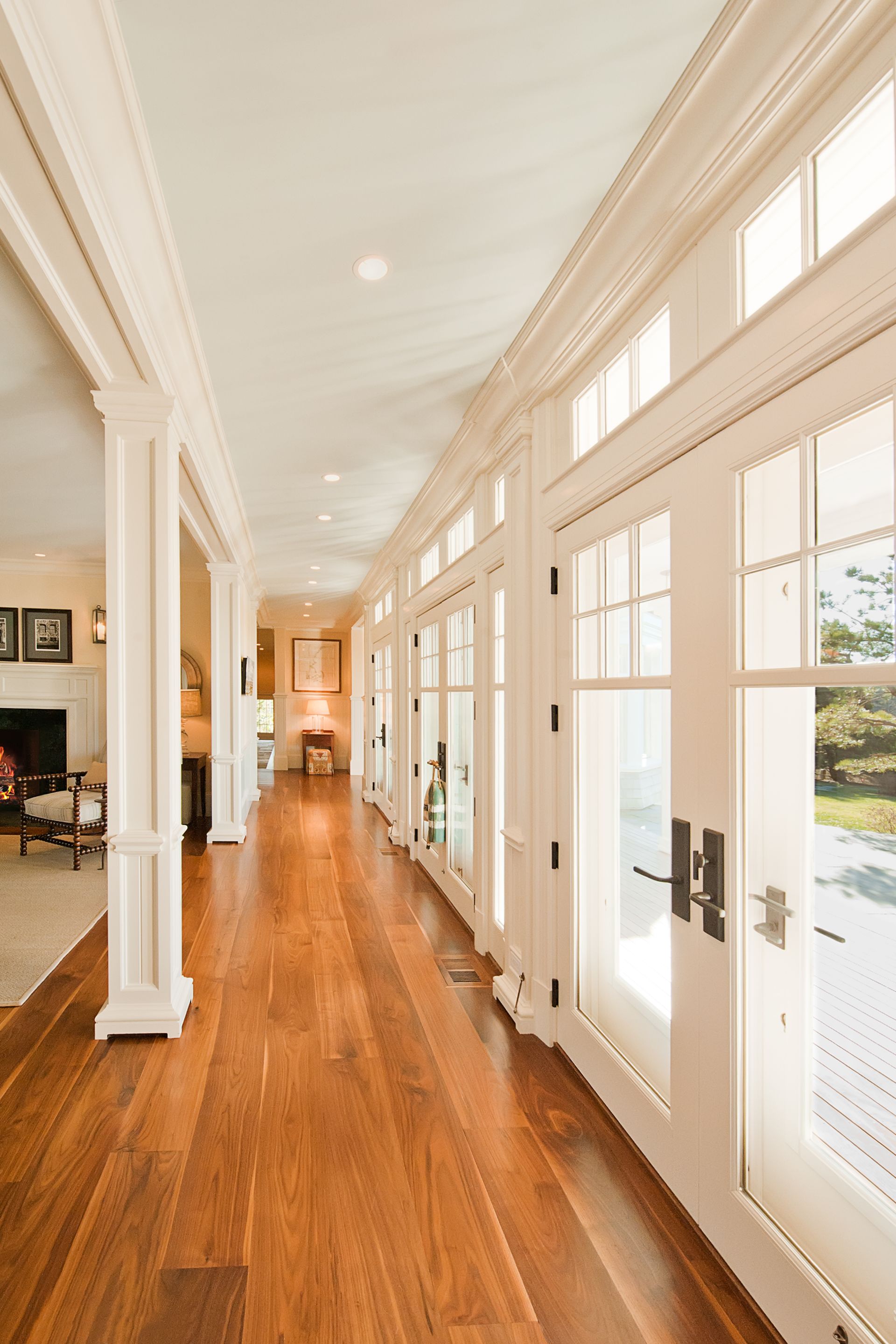 A long, bright hallway with polished wood floors, white paneled walls, and a series of glass doors leading outside.