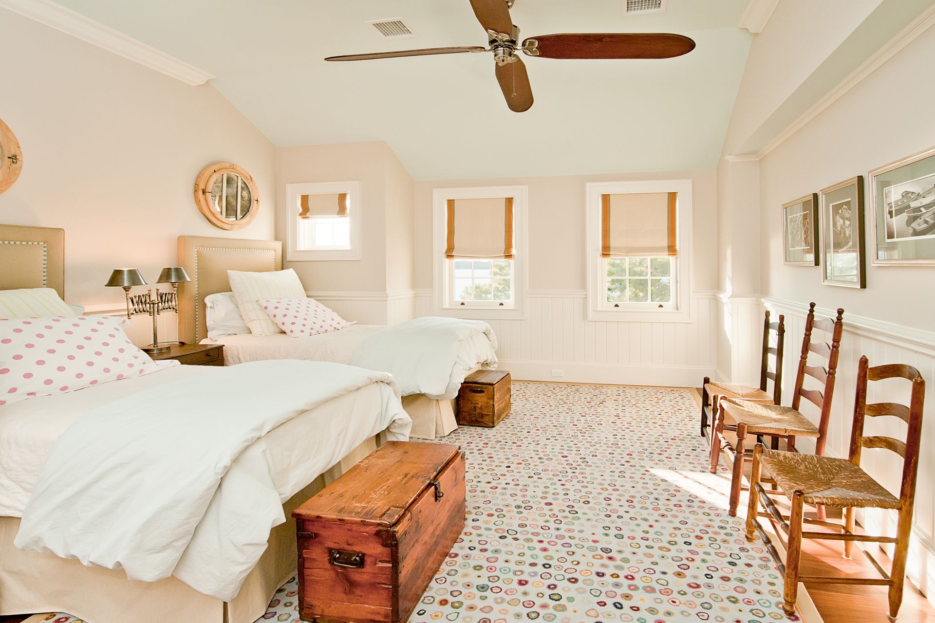 A bedroom with two beds, wooden chests, a ceiling fan, and three wooden chairs lined up against a wall with artwork.