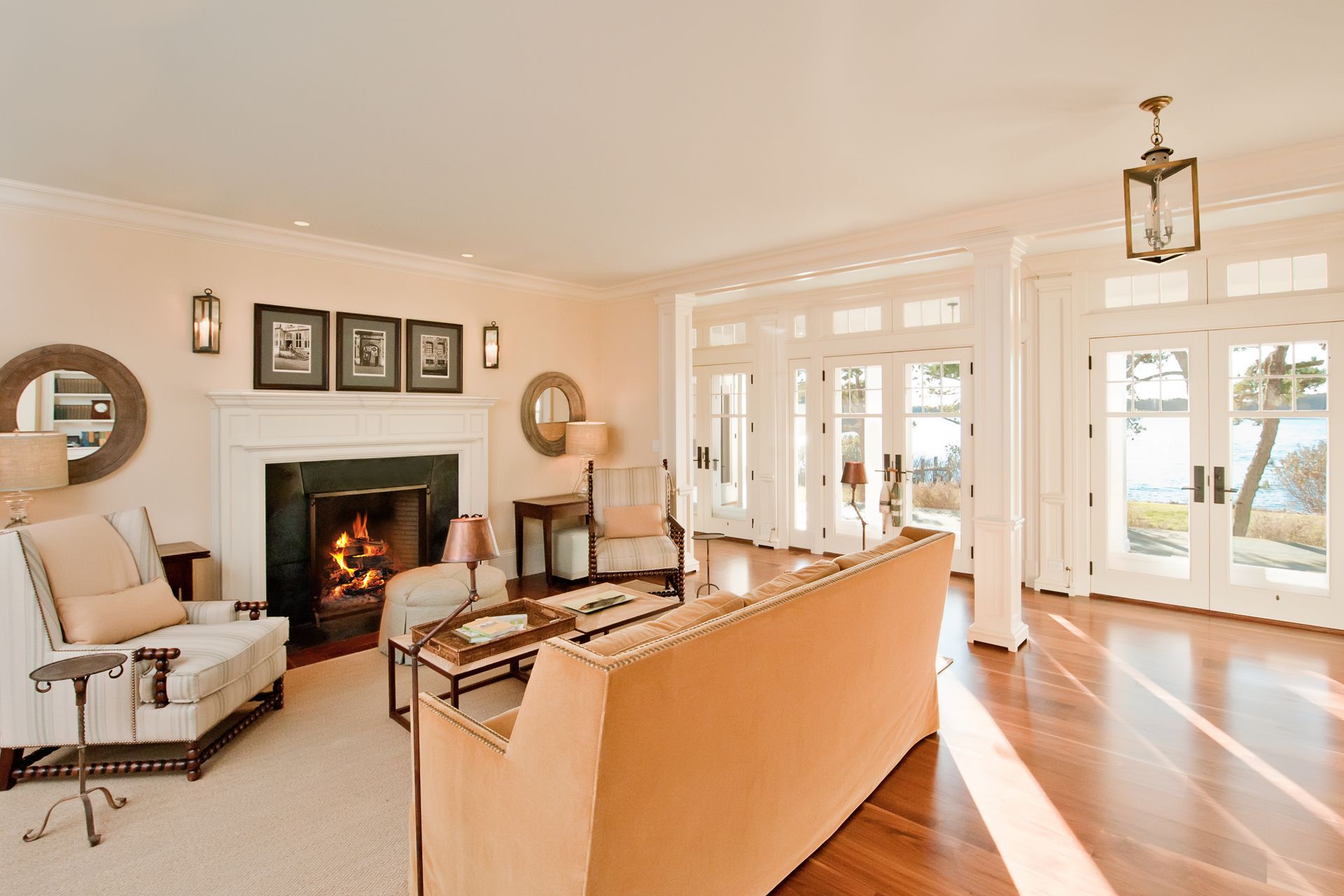 A bright living room with a fireplace, sofa, armchair, and floor-to-ceiling glass doors overlooking a waterfront view.