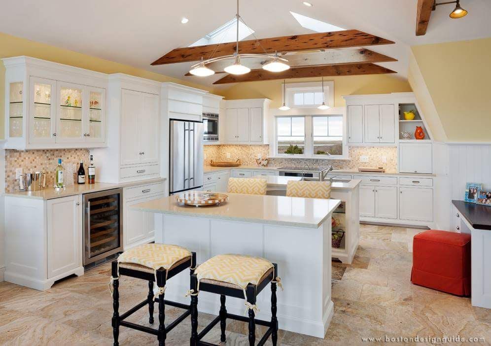 Bright, modern kitchen with white cabinetry, a central island with two bar stools, wooden beams, and stone tile flooring.